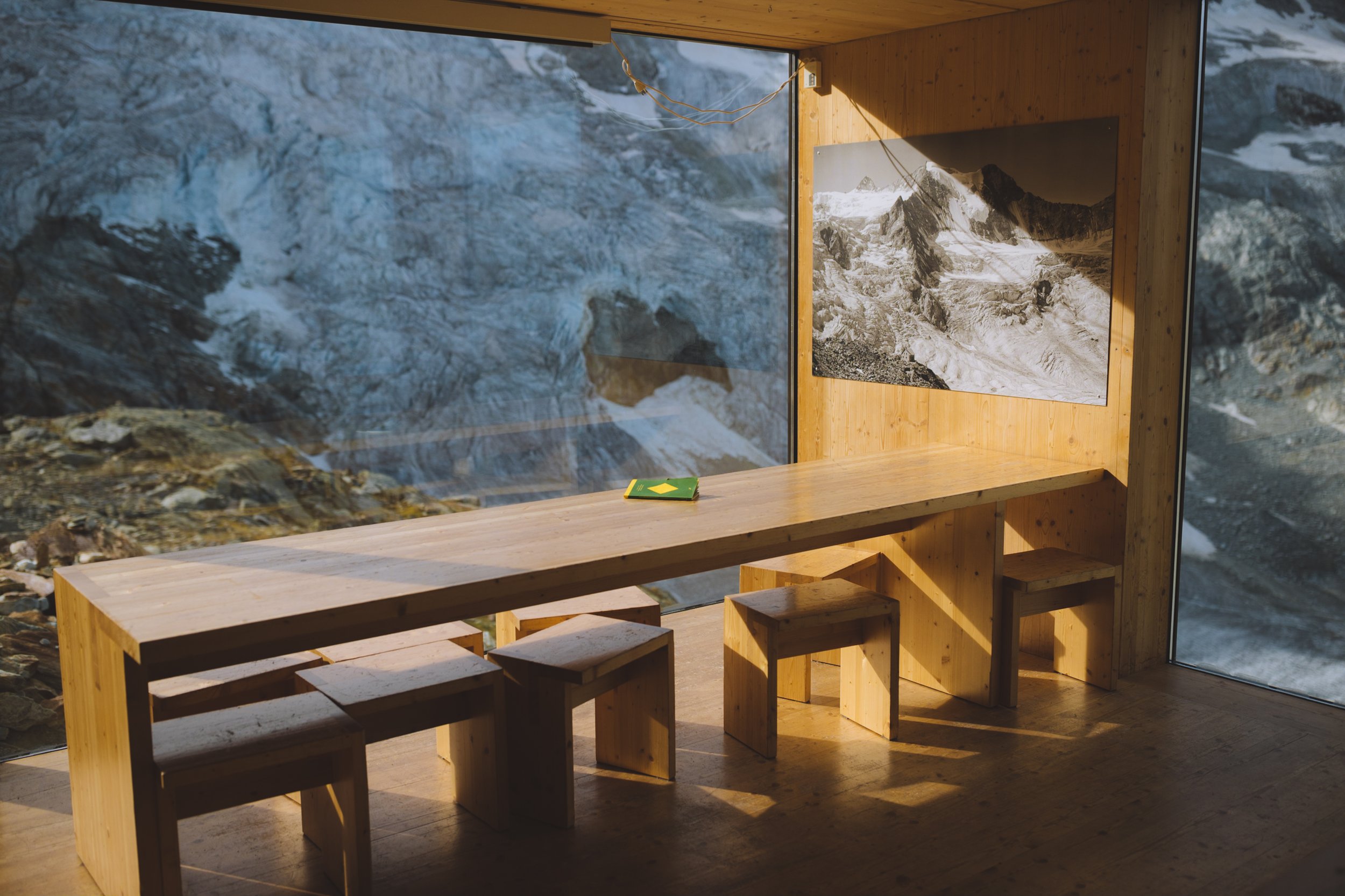A long wooden table with wooden and wooden chair's inside a building with a large glass window looking out onto a rocky mountainside along the Haute route in Switzerland