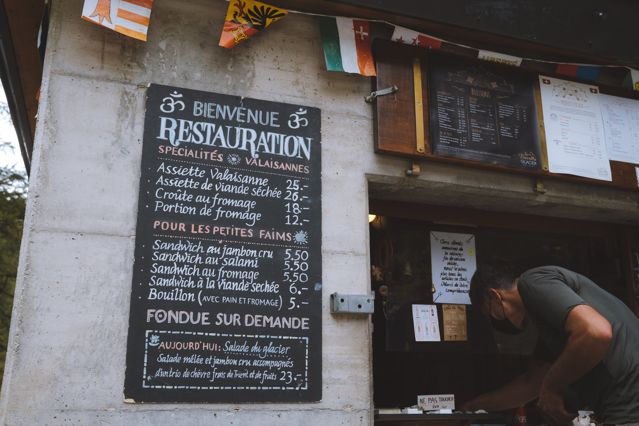 A small concrete restaurant with a black and white menu outside listing the food available in Swiss French