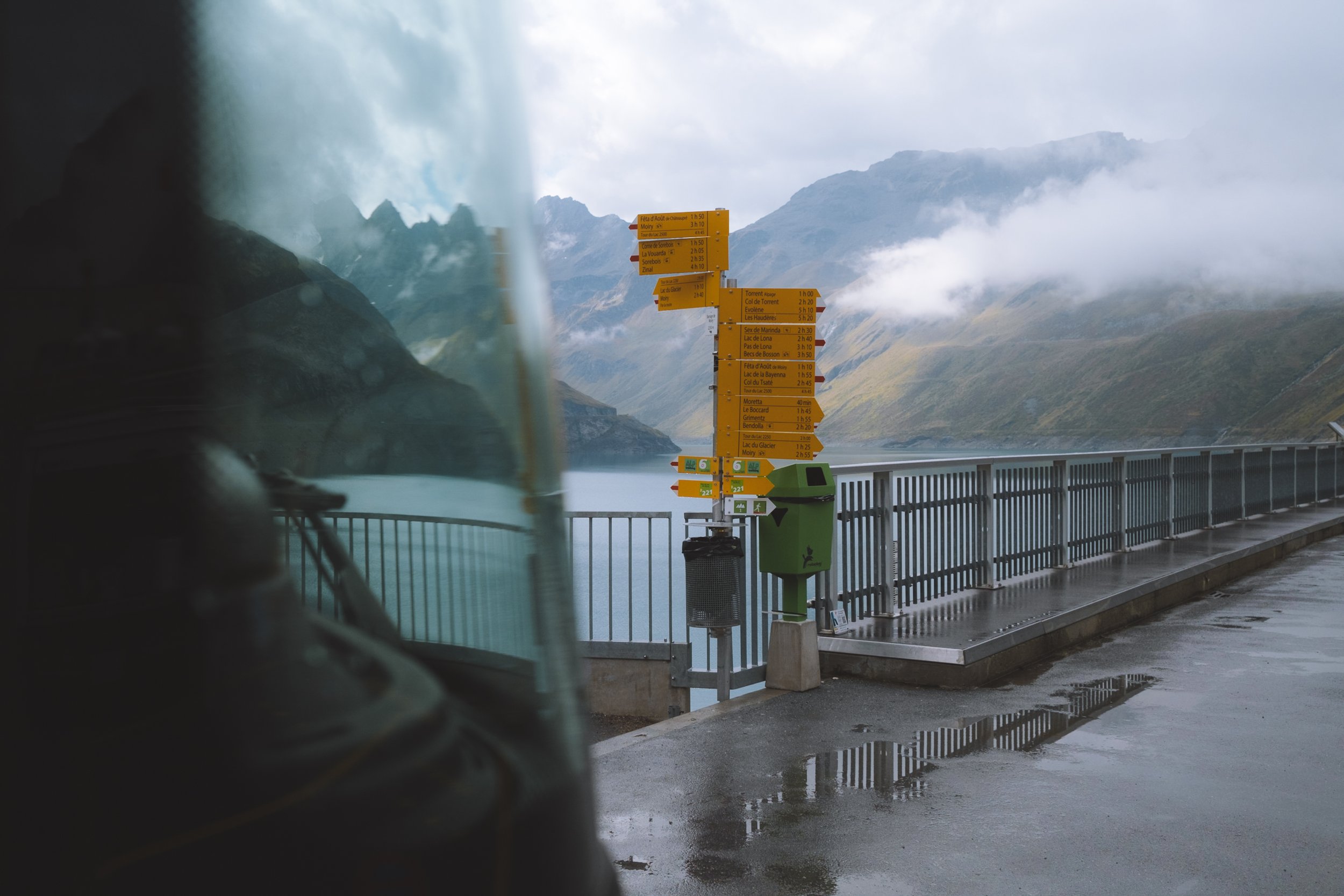 Standing next to a bus window looking at a post with many yellow signs at the edge of a railing with a large body of water and mountains in the distance 