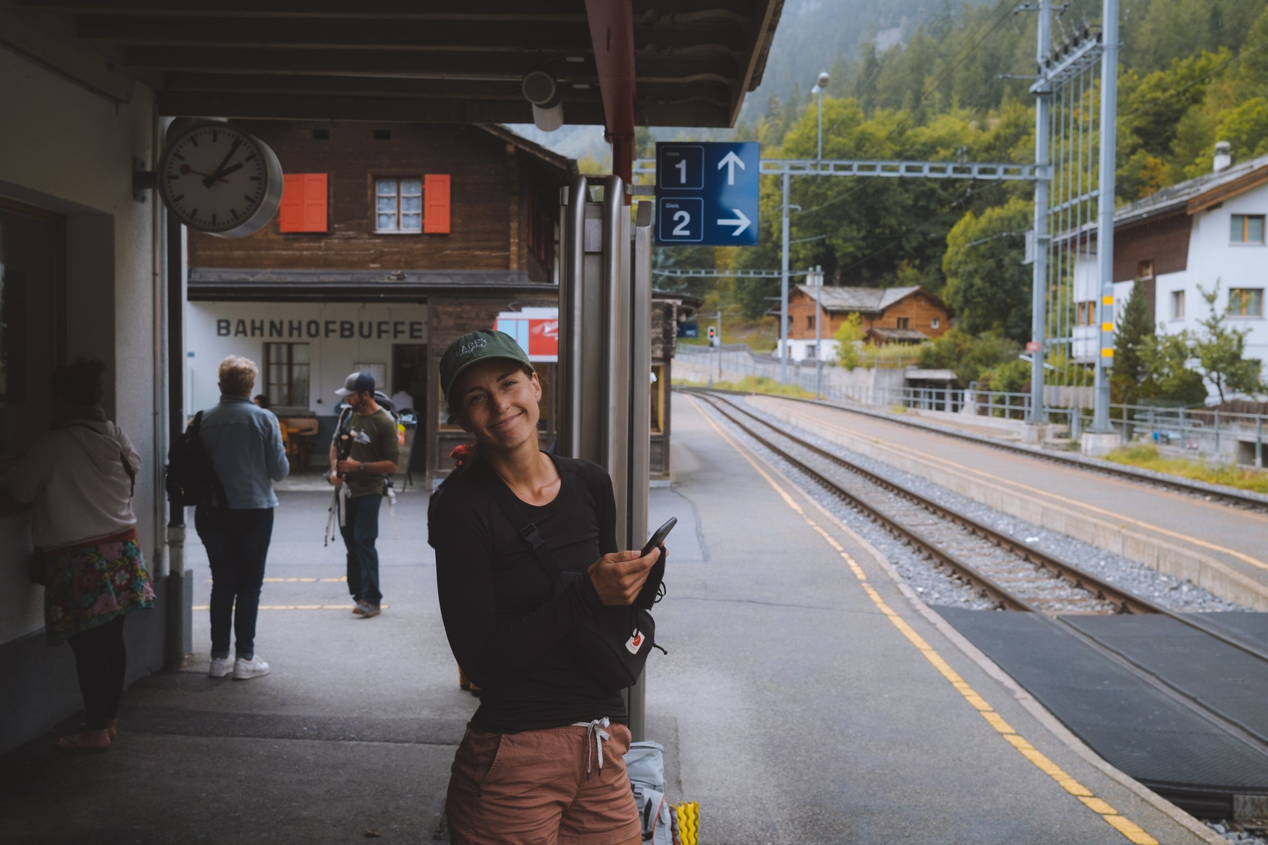 Andrea in black standing at a train station platform with the train tracks to her right and other people waiting for the train on her left 